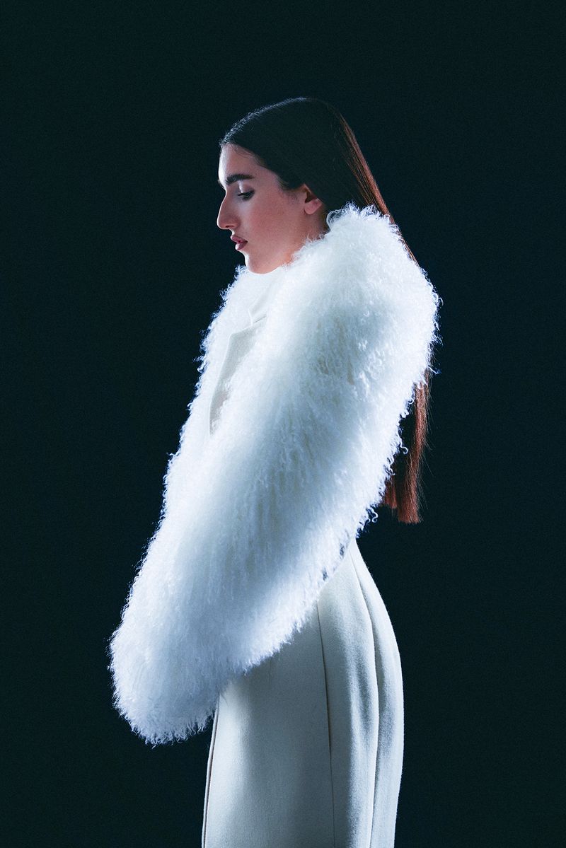 Elegant portrait of a woman in profile wearing a white coat with a fur collar against a black background in a studio setting.
