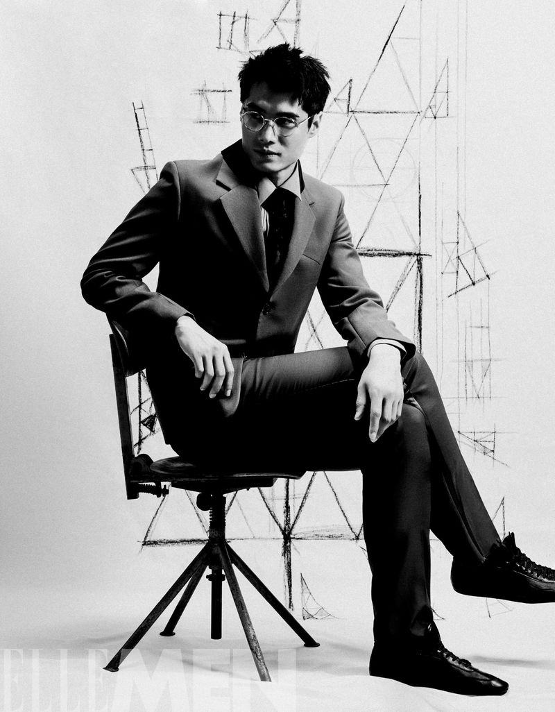 Black and white portrait of a stylish adult male in a suit, seated on a chair, with geometric line art in the background.