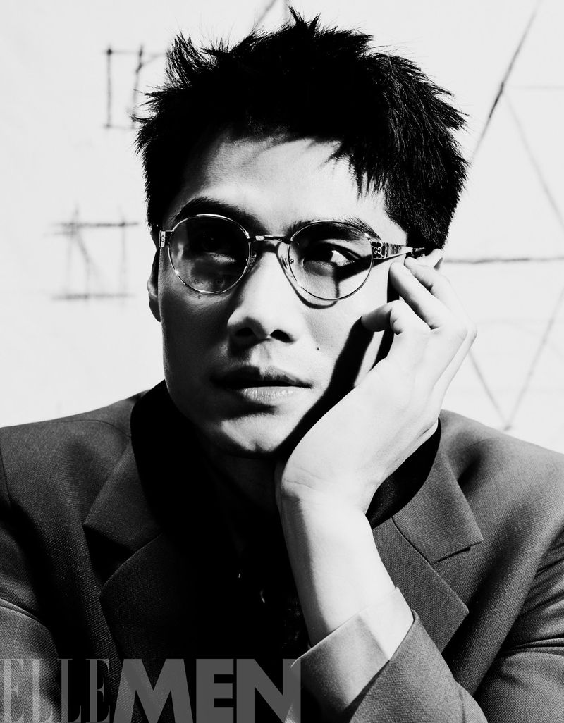 Black and white portrait of a stylish adult male with glasses and a dark suit, in a studio setting with sketched background.