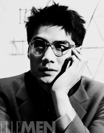 Black and white portrait of a stylish adult male with glasses and a dark suit, in a studio setting with sketched background.