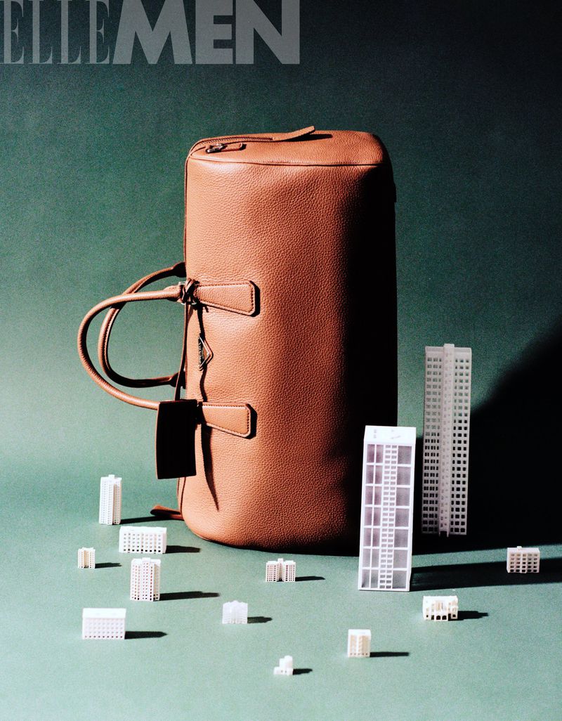 Studio shot of a brown leather duffel bag with miniature building models on a green surface, casting shadows, for ELLE MEN magazine.