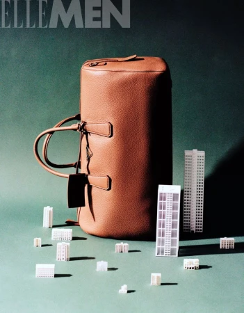 Studio shot of a brown leather duffel bag with miniature building models on a green surface, casting shadows, for ELLE MEN magazine.