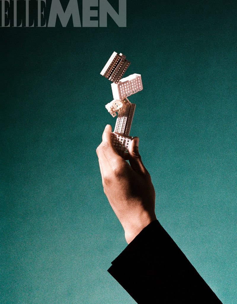 Hand holds a tower of building blocks against a teal background. Fashionable black jacket sleeve is visible in the lower right corner.