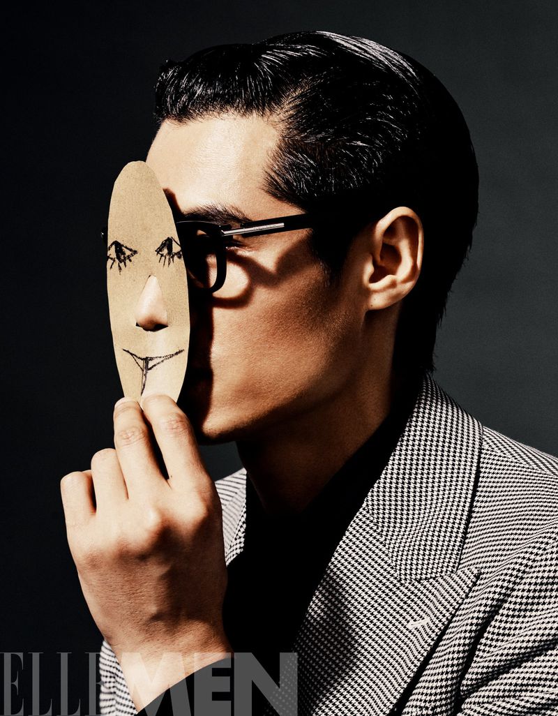 Stylish portrait of a man in a blazer and glasses holding a drawn face in front of his face in an indoor setting with studio lighting.