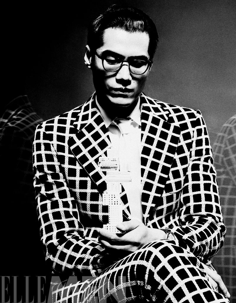 Black and white portrait of a man wearing glasses and a patterned suit, looking down with a serious expression, with dark background.