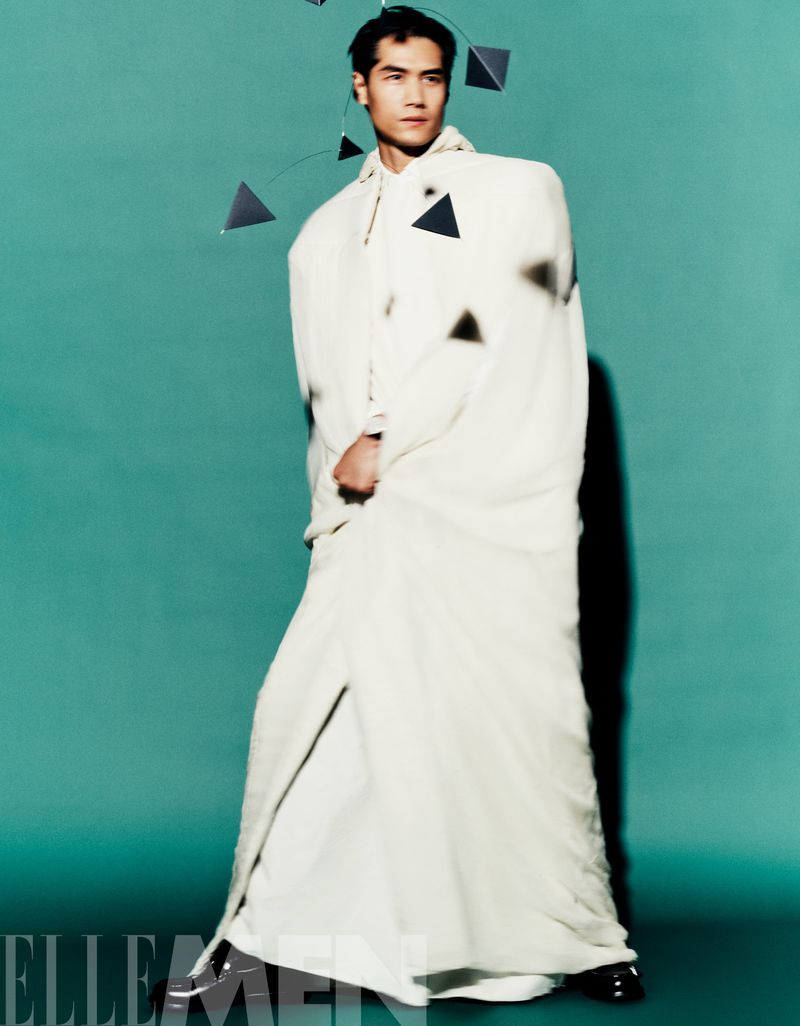 Elegant Asian male model poses in a full-length shot wearing an off-white robe, dress shoes, and a geometric sculpture near his head.