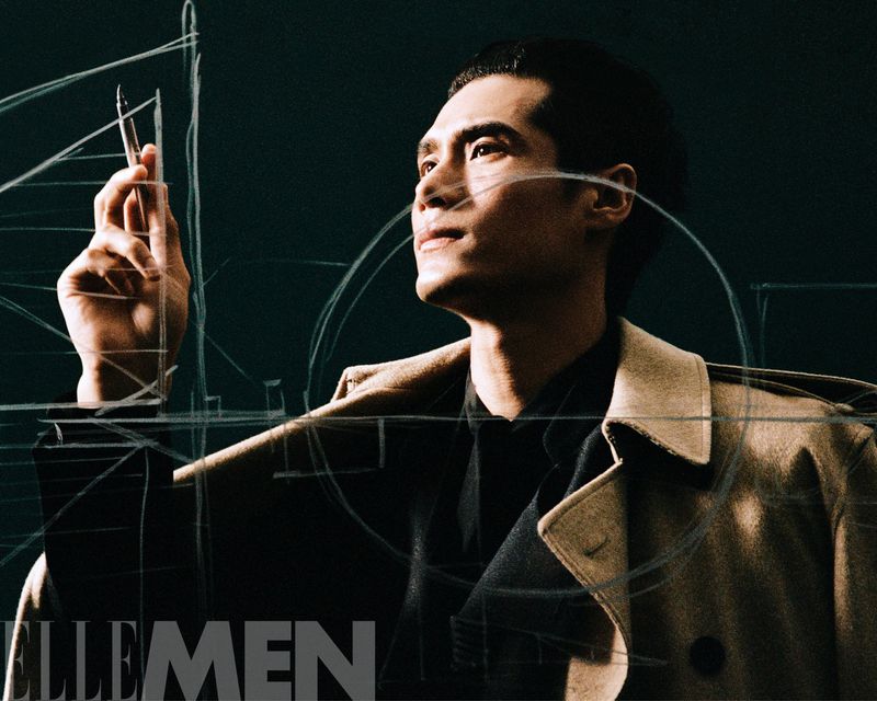 Portrait of a dark-haired Asian man in a trench coat, holding a drawing tool, with geometric lines overlaid and the Elle Men logo.