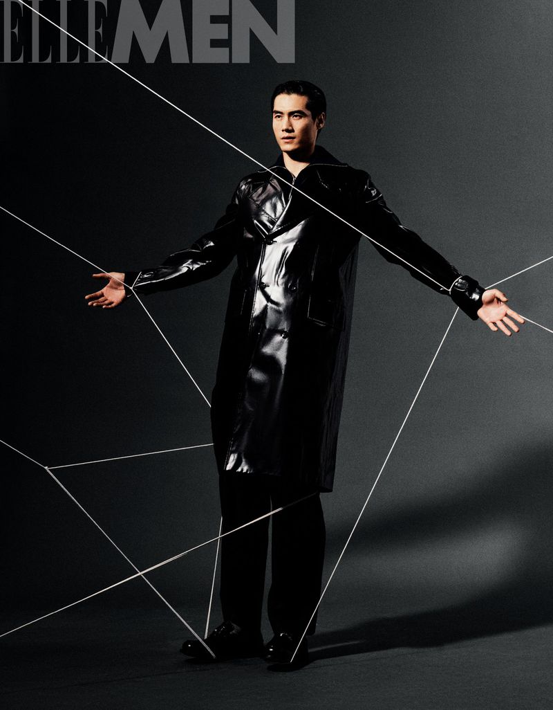 Editorial shot of adult male model in glossy black coat with minimalist lighting and geometric string art against a dark gray background.