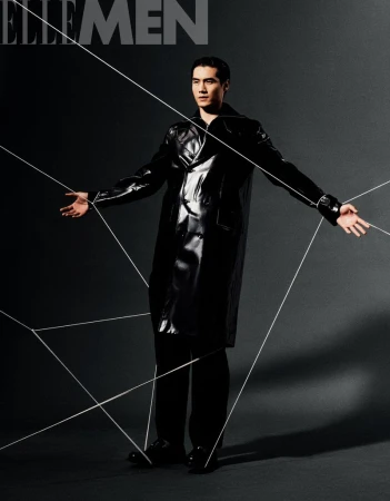 Editorial shot of adult male model in glossy black coat with minimalist lighting and geometric string art against a dark gray background.