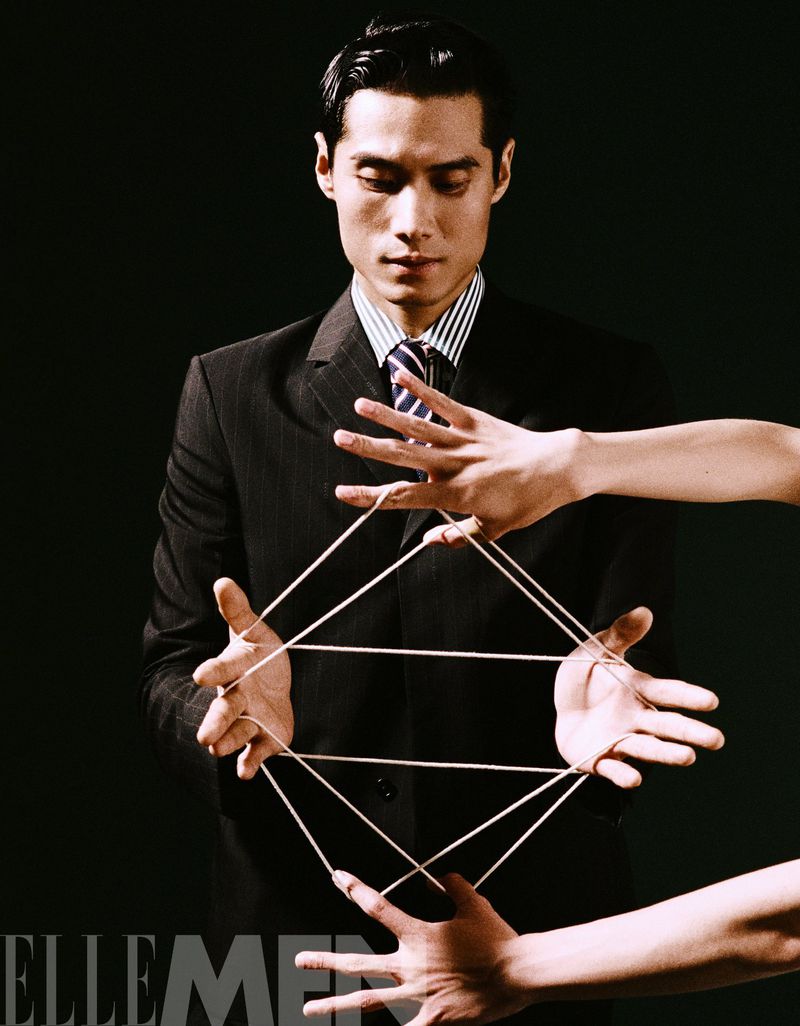 Elegant Asian man in suit performs string figure with disembodied hands against black background. Conceptual fashion advertisement.