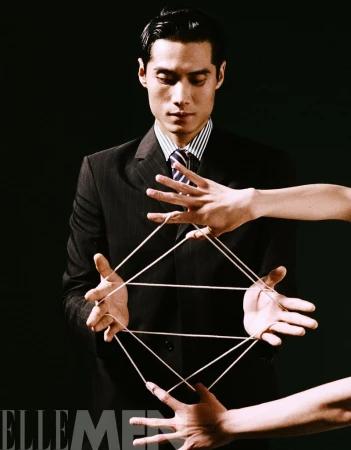 Elegant Asian man in suit performs string figure with disembodied hands against black background. Conceptual fashion advertisement.