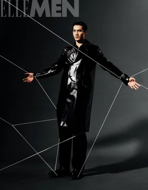 Model wearing a black glossy trench coat on a dark grey seamless studio backdrop intersected by thin white strings.
