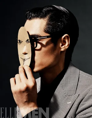 Model holding a small cutout cardboard mask with a drawn face in front of his eye, wearing a houndstooth blazer.