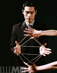 Asian man in a dark suit holding a complex geometric cat's cradle string figure against a black background.