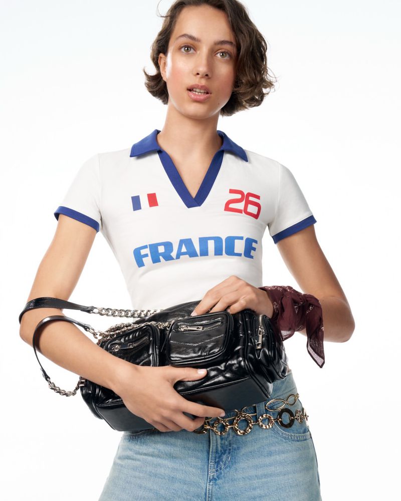 Model posing in a white France polo shirt and denim jeans holding a glossy black multi-pocket shoulder bag against a white background.