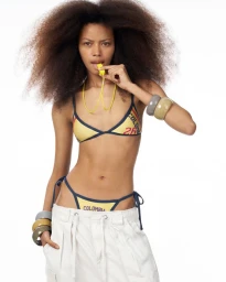 Janine Suarez poses in a yellow Colombia bikini and white cargo pants with a yellow whistle in front of a white studio backdrop.