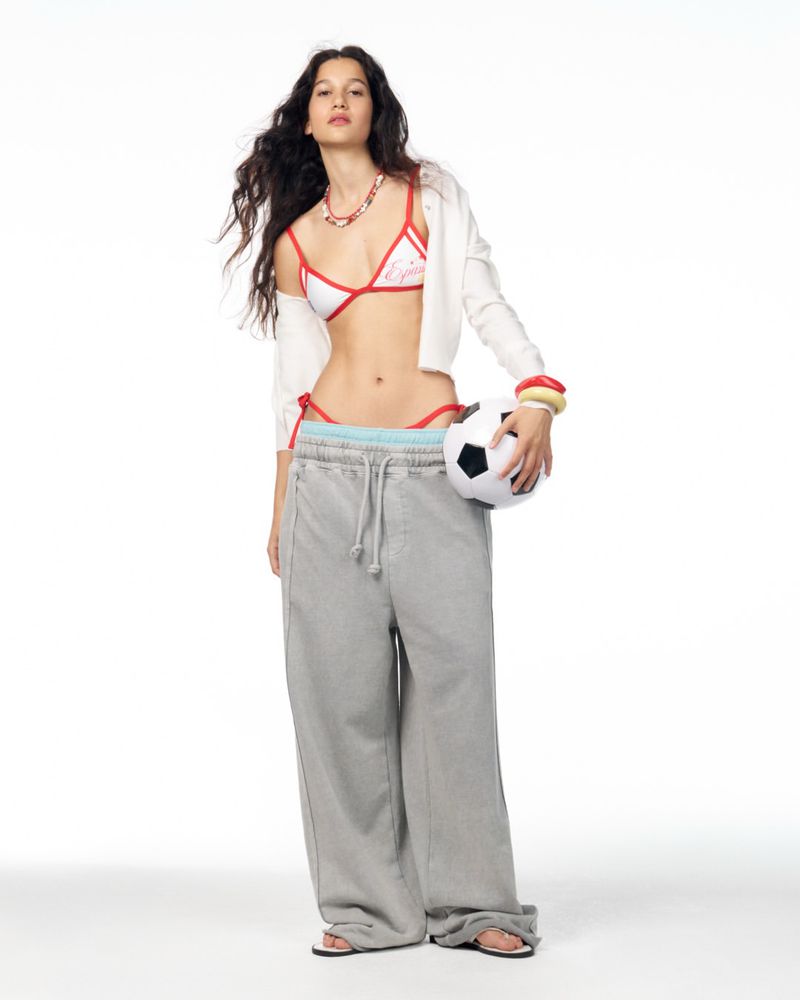 Model Janine Suarez in oversized grey sweatpants and a white bikini top holding a soccer ball against a white studio backdrop.