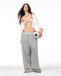 Model Janine Suarez in oversized grey sweatpants and a white bikini top holding a soccer ball against a white studio backdrop.
