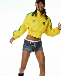 Model posing in a yellow cropped athletic jacket with green trim and denim cutoff shorts against a white backdrop.