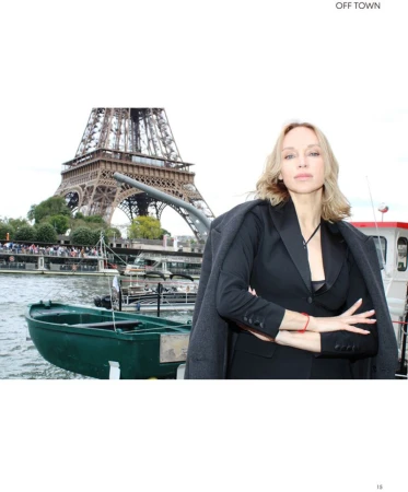 Adult woman in a black blazer standing in front of the Eiffel Tower in Paris, with a boat on the river Seine.