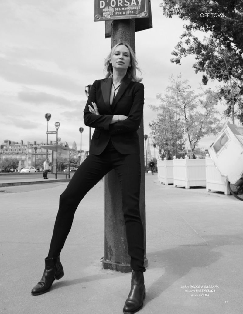 Blonde woman in black suit and boots poses in urban street scene in black and white. Street sign and building in background.