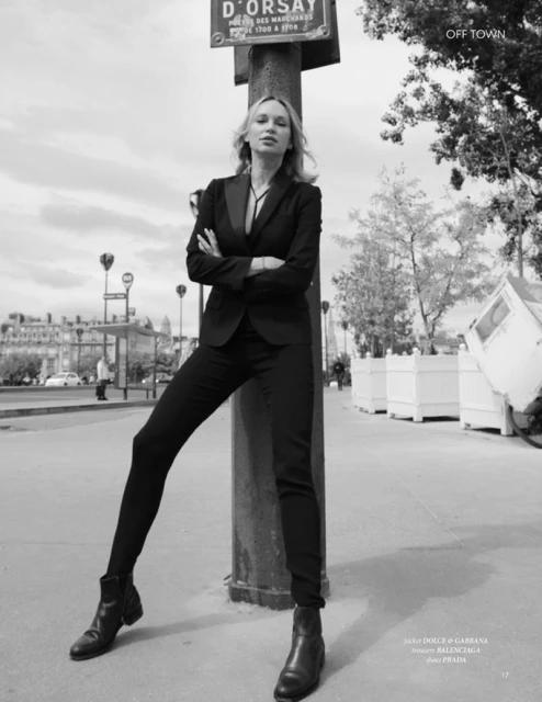 Blonde woman in black suit and boots poses in urban street scene in black and white. Street sign and building in background.