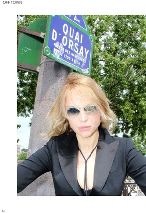 Model wearing a black blazer and sunglasses in front of a blue Quai d Orsay street sign and trees in Paris.