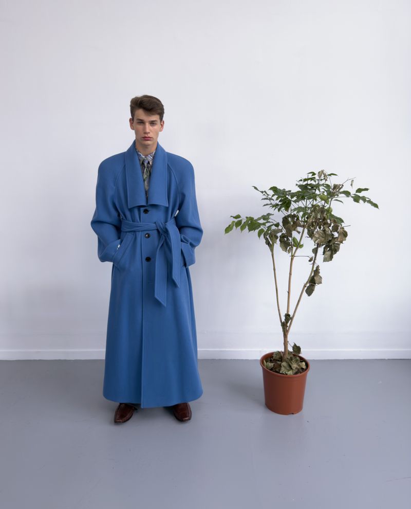 A young male model stands in a full-length belted blue coat next to a potted houseplant against a plain white wall and grey studio floor.