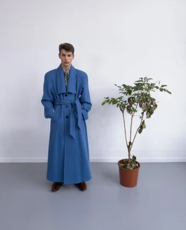 A young male model stands in a full-length belted blue coat next to a potted houseplant against a plain white wall and grey studio floor.