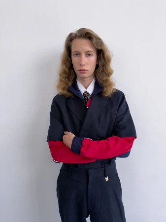 Male model with long wavy hair posing with arms crossed in a navy suit with red sleeves against a plain white backdrop.