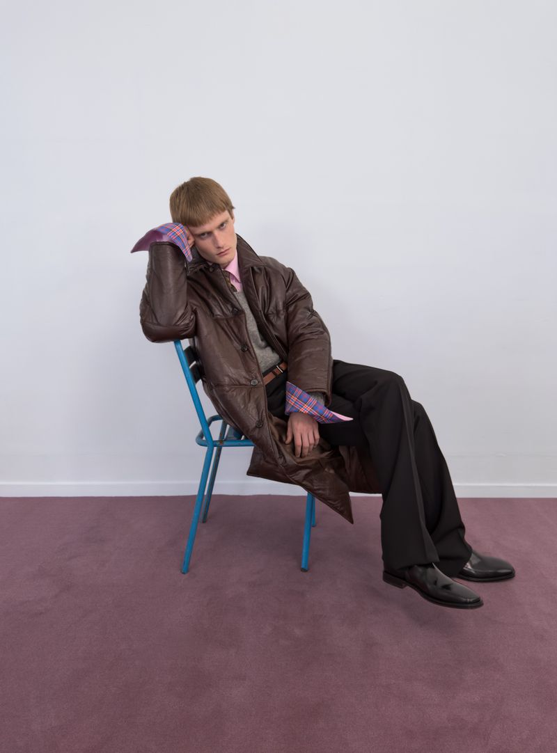 Male model seated on a blue metal chair wearing a long brown quilted coat and black trousers against a white wall and mauve carpet.