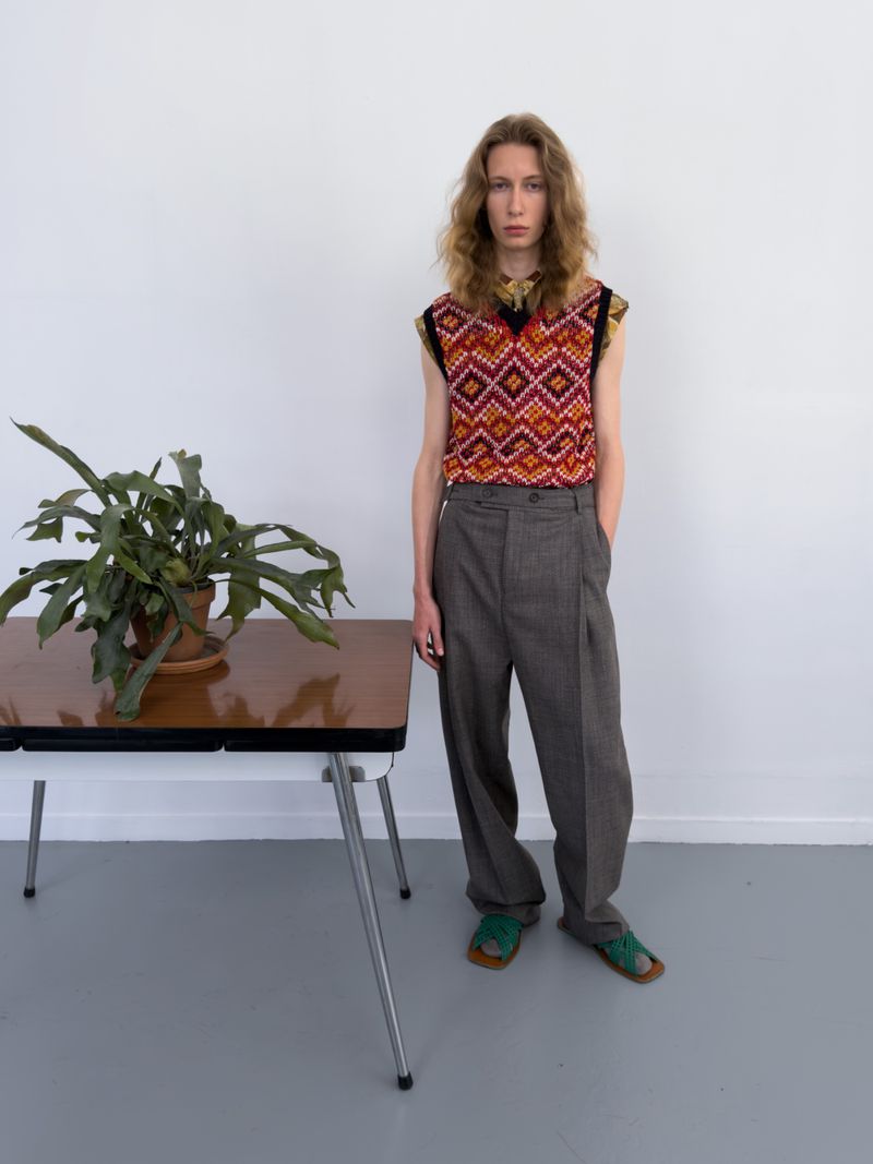 Model standing next to a potted plant on a wooden desk wearing a geometric knit sweater vest, pleated grey trousers, and green sandals.