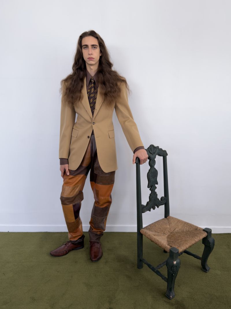 Model with long brown hair wearing a tan blazer and patchwork leather trousers standing beside an ornate chair on olive green carpet.