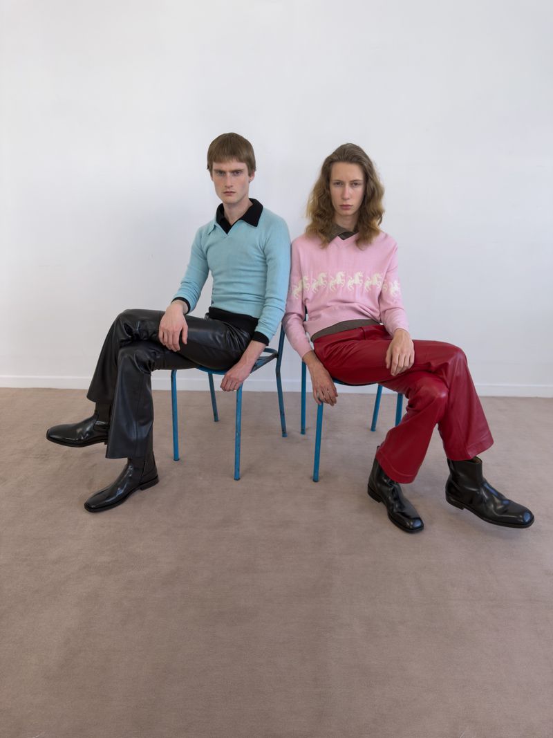 Two male models seated side by side on blue chairs wearing a light blue polo and a pink sweater with red and black leather pants.