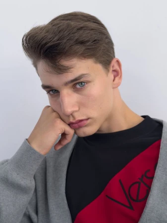 Portrait of a young man resting his head on his hand wearing a grey cardigan and black top against a white studio background.