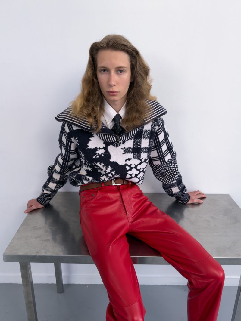 A model wearing red leather pants and a black and white patterned knit zip sweater seated on a metal table against a white wall.
