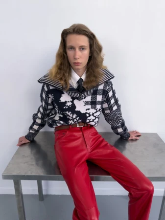 A model wearing red leather pants and a black and white patterned knit zip sweater seated on a metal table against a white wall.