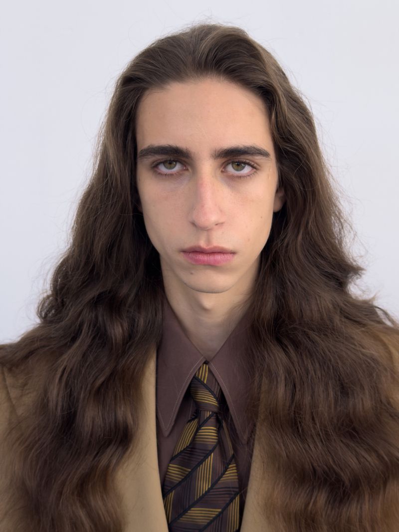 Portrait of a man with long brown hair wearing a brown suit jacket, shirt, and patterned tie against a plain white background.