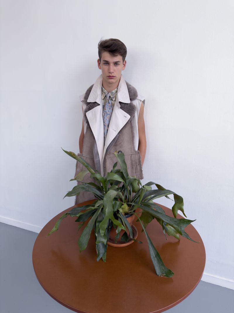 Young male model in a sleeveless shearling vest standing behind a round brown table with a potted plant against a white wall.
