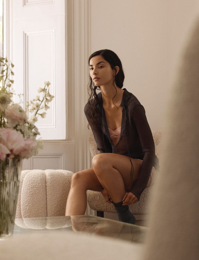 Woman in casual outfit sits near a window in a light-filled room, adjusting her sock, with flowers and furniture visible.