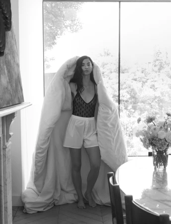 Young woman in lingerie and shorts stands indoors by a window, partially enveloped in a white duvet, with a marble table and flowers nearby.