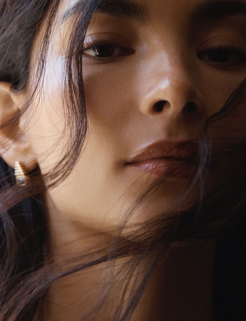 Close-up portrait of a woman with brown hair and neutral makeup, featuring a gold earring, captured in a soft, diffused lighting.