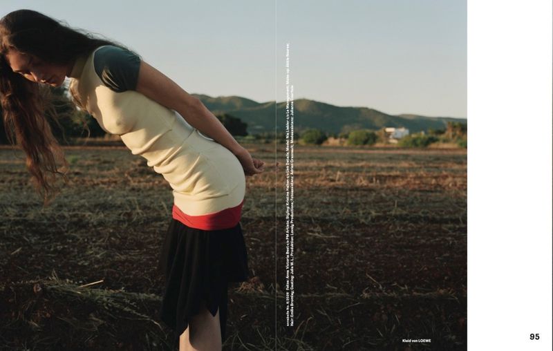 Model Nia Liebler bends forward wearing a fitted cream and black dress in a dry rural field against distant rolling hills.