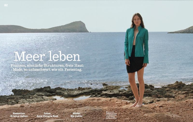Model Nia Liebler poses barefoot on a rocky shore wearing a teal button-up shirt and black skirt against an ocean backdrop.