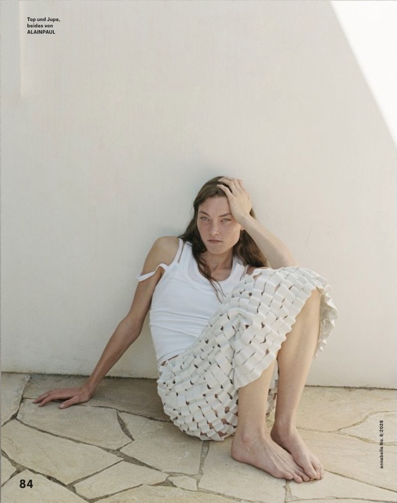 Model Nia Liebler sitting barefoot on a flagstone floor against a white wall, wearing a white tank top and a woven lattice skirt.
