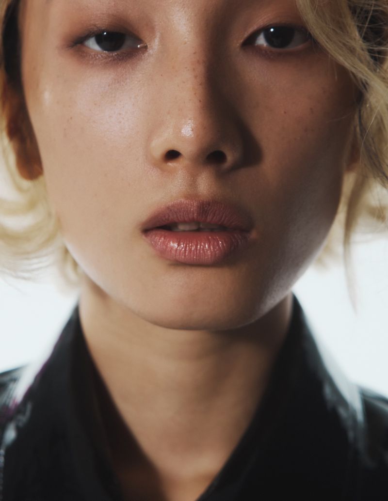 Close-up beauty portrait of an East Asian model with blonde hair and freckles against a bright background.
