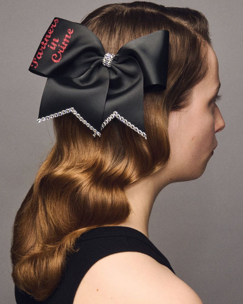 Adult female profile wearing a large black hair bow with 'Partners in Crime' inscription and hair styled in vintage waves, in studio.
