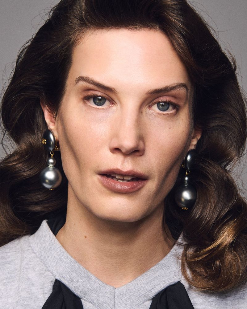 Close-up of a brunette adult female looking at the camera wearing large, gray ball drop earrings against a gray backdrop.