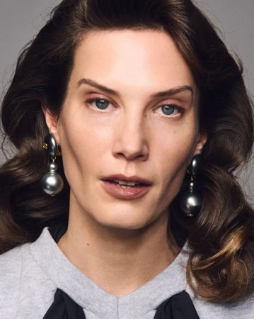 Close-up of a brunette adult female looking at the camera wearing large, gray ball drop earrings against a gray backdrop.
