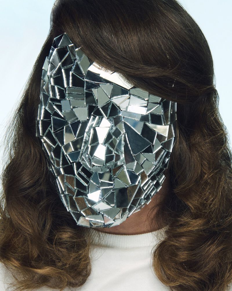 Person wearing a reflective mirrored mask covering their face, with long brown hair. The mask consists of triangular and square mirrors.